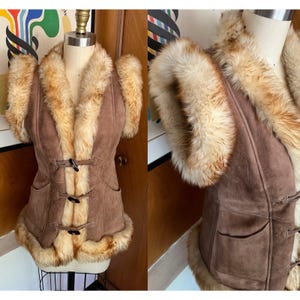 May include: A brown suede vest with a fur collar and trim. The vest has a toggle closure and two front pockets. The fur is a light brown color. The vest is displayed on a mannequin.
