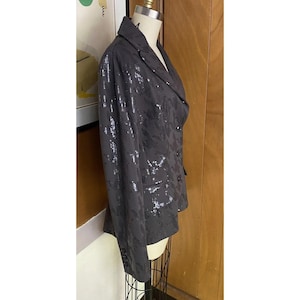 K&ouml;nnte beinhalten: Ein schwarzer Blazer mit einem silbernen Paillettenmuster. Der Blazer hat einen Schalkragen, lange &Auml;rmel und einen zweireihigen Verschluss.