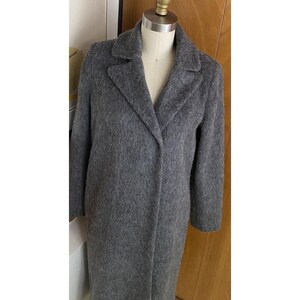 Op de afbeelding: Een lange, grijze wollen jas met een klassieke reverskraag. De jas wordt getoond op een mannequin, die de getailleerde pasvorm en lengte laat zien. De kleur van de jas is een middengrijs en de stof lijkt een zachte textuur te hebben.
