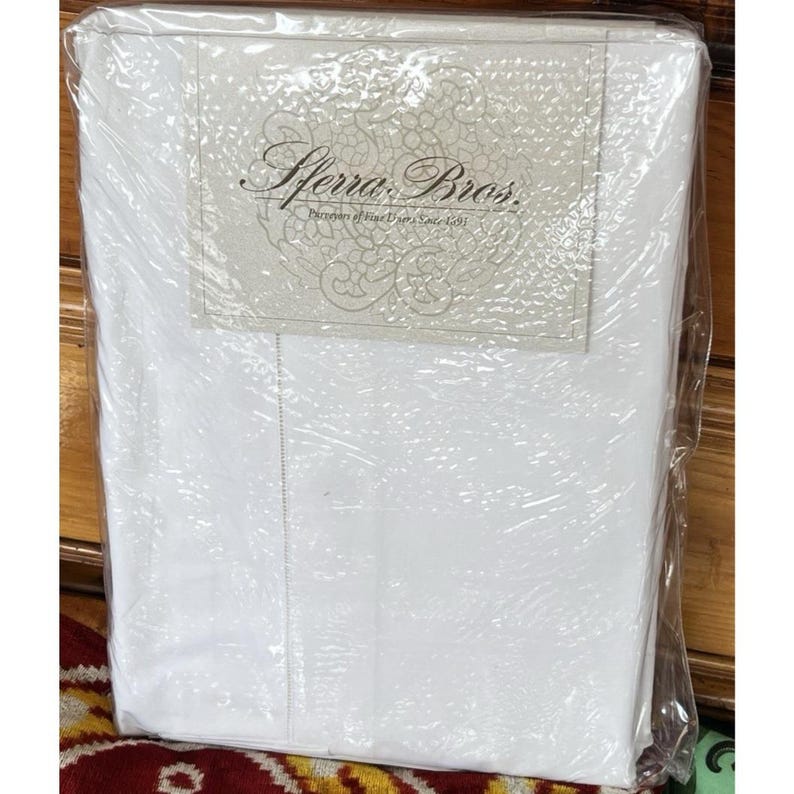 Pu&ograve; includere: Un set di lenzuola bianche confezionato. La confezione include un'etichetta con la scritta "Sferra Bros. Purveyors of Fine Linens Since 1891". Le lenzuola sono visibili attraverso l'involucro trasparente.