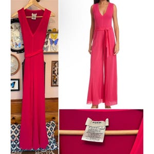 May include: A sleeveless, fuchsia jumpsuit with a V-neckline and wide legs. The jumpsuit features a matching fabric belt that cinches the waist. The brand label "Fuzzi" is visible. The garment is displayed on a wooden hanger.