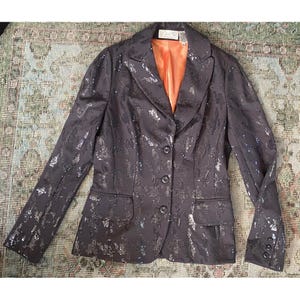 K&ouml;nnte beinhalten: Ein schwarzer Blazer mit silbernen Pailletten, mit einem einzigen Knopfverschluss und einer Tasche auf der linken Seite. Der Blazer hat ein Satin-Futter in einer verbrannten Oranget&ouml;nung.