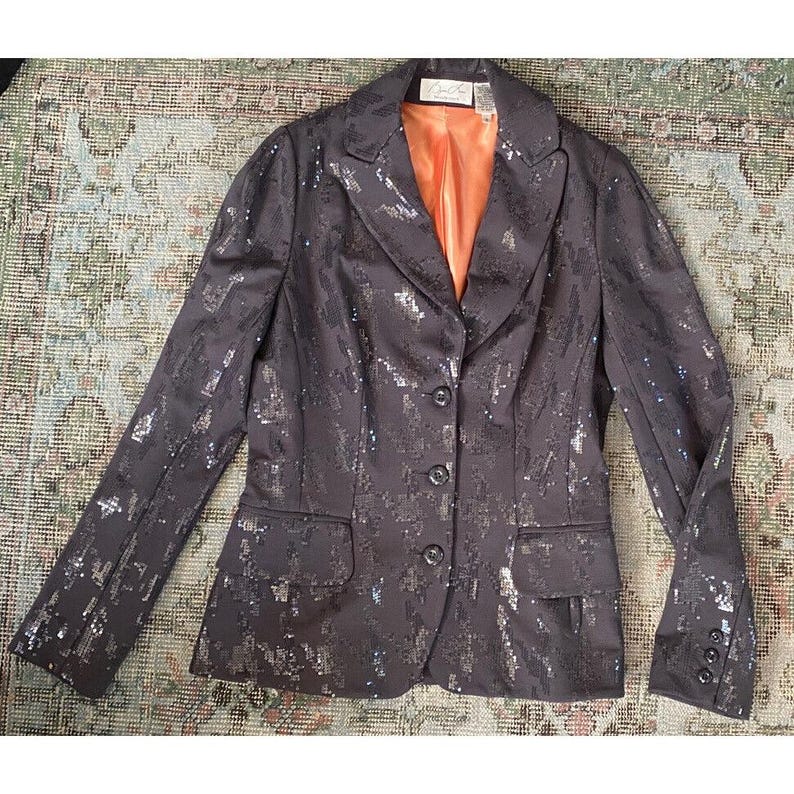 K&ouml;nnte beinhalten: Ein schwarzer Blazer mit silbernen Pailletten, mit einem einzigen Knopfverschluss und einer Tasche auf der linken Seite. Der Blazer hat ein Satin-Futter in einer verbrannten Oranget&ouml;nung.