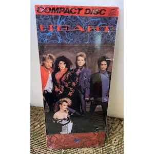 May include: A compact disc case featuring the band Heart. The cover has the words "COMPACT DISC" at the top and "HEART" in large letters. The image shows the band members in 1980s attire, with a dark background.