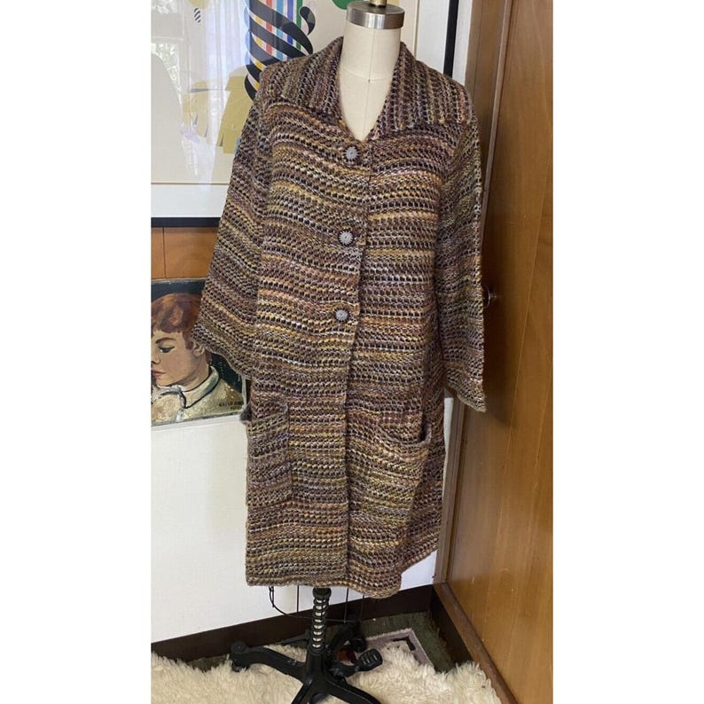 K&ouml;nnte beinhalten: Ein Vintage-Mantel mit langen &Auml;rmeln in einem gewebten Muster aus Braun, Beige und Lila. Der Mantel hat einen Kragen, drei gro&szlig;e Kn&ouml;pfe und zwei Vordertaschen. Der Mantel wird auf einer Schaufensterpuppe pr&auml;sentiert.