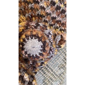 K&ouml;nnte beinhalten: Nahaufnahme eines handgefertigten, strukturierten, mehrfarbigen Strickartikels. Der Artikel hat ein zentrales, kreisf&ouml;rmiges Design mit einem grauen Mittelteil und braunen und schwarzen Akzenten. Das umgebende Strickgewebe hat eine Mischung aus braunen, gelben und schwarzen Farbt&ouml;nen.