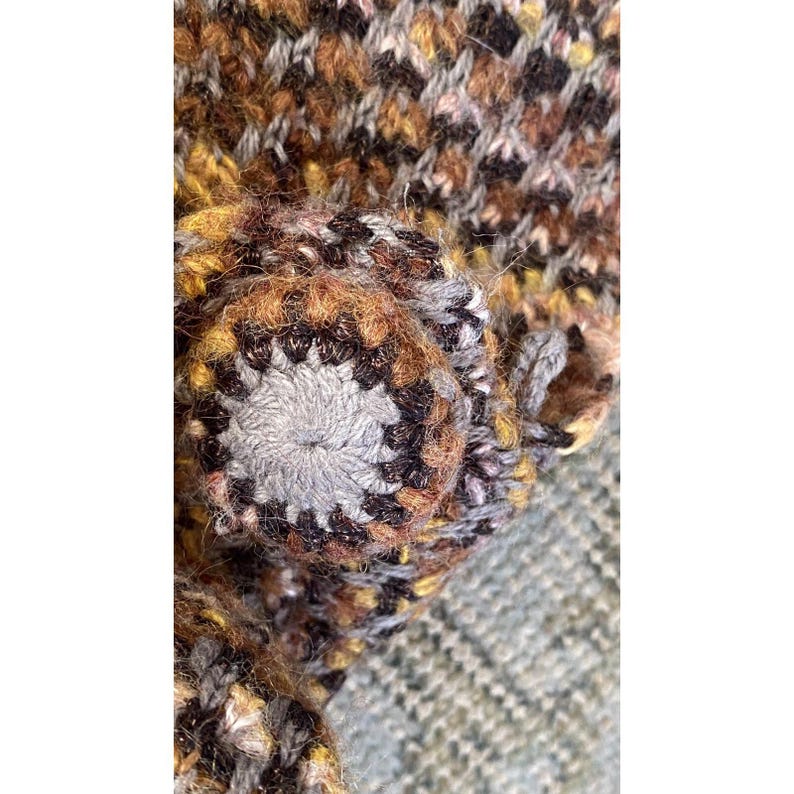 K&ouml;nnte beinhalten: Nahaufnahme eines handgefertigten, strukturierten, mehrfarbigen Strickartikels. Der Artikel hat ein zentrales, kreisf&ouml;rmiges Design mit einem grauen Mittelteil und braunen und schwarzen Akzenten. Das umgebende Strickgewebe hat eine Mischung aus braunen, gelben und schwarzen Farbt&ouml;nen.