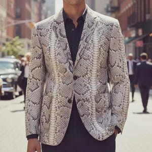Pode incluir: Um blazer com padrão de pele de cobra em tons de cinza e branco. O casaco tem lapelas entalhadas, dois botões e dois bolsos frontais. A pessoa está usando uma camisa preta por baixo. O fundo mostra uma rua da cidade.