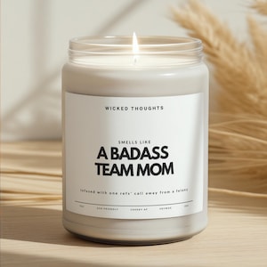 May include: A lit, white soy wax candle in a clear glass jar. The label reads "WICKED THOUGHTS" and "SMELLS LIKE A BADASS TEAM MOM." The text "Infused with one refs' call away from a felony" is also on the label. The candle is eco-friendly.