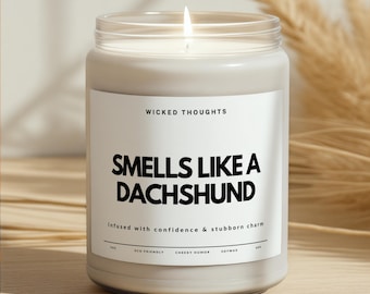 May include: A lit, white candle in a clear glass jar. The label reads "WICKED THOUGHTS SMELLS LIKE A DACHSHUND" with the text "Infused with confidence & stubborn charm". The candle is eco-friendly and made with soy wax.