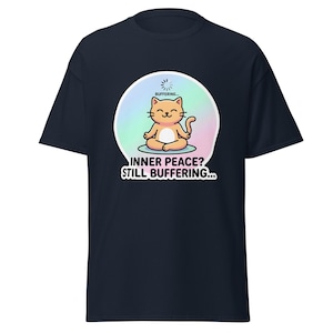 Può includere: T-shirt blu navy con un gatto dei cartoni animati che medita. Il gatto arancione è in posizione del loto. Il testo recita "INNER PEACE? STILL BUFFERING..." e "BUFFERING...". Il design è su uno sfondo dai colori pastello.