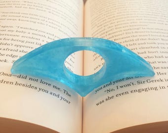 May include: A light blue resin thumb page holder with a circular opening, resting on an open book. The page holder is shaped like a stylized wing. The book's pages are yellowed with text.