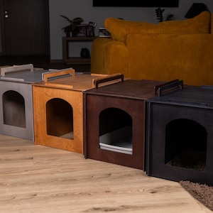 May include: A row of five wooden cat litter box enclosures in various colors: beige, gray, light brown, dark brown, and black. Each box has an arched entrance and a handle on top. The boxes are on a wooden floor.