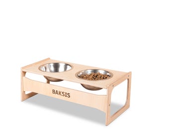 May include: A light-colored wooden pet feeder with two stainless steel bowls. One bowl is filled with dry pet food. The word "BAKSIS" is engraved on the front of the feeder.