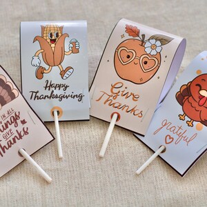 Puede incluir: Cuatro portachupetes con temática de Acción de Gracias con ilustraciones de dibujos animados. Uno presenta un pavo, otro una manzana con gafas de corazón, un personaje de maíz y un pavo. Cada uno tiene un palito de chupete blanco y texto festivo.