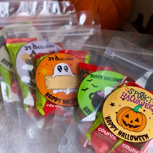Può includere: Sacchetti di plastica trasparente pieni di dolcetti a tema Halloween. Ogni sacchetto ha un adesivo circolare con grafica e testo di Halloween, tra cui fantasmi, zucche e le parole "Happy Halloween" e "Spooky Halloween."