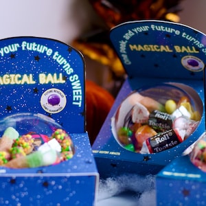 Op de afbeelding: Blauwe dozen met de tekst "MAGICAL BALL" bevatten snoepjes onder een transparante koepel. De dozen zijn versierd met sterren en de tekst "One peek, and your future turns sweet."