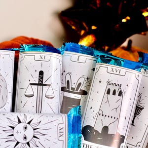 May include: White tarot card designs with silver and blue foil wrapping. The cards feature illustrations of a lion, scales, a magician's hat, a tower, and a sun. The cards are displayed with pumpkins in the background.