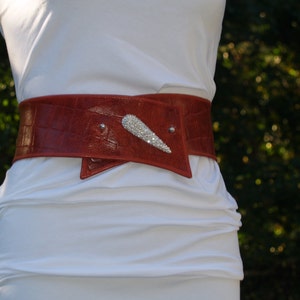 May include: A red leather belt with a decorative silver buckle. The belt is worn over a white shirt.