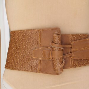 May include: A wide, tan-colored belt with a woven texture on the front panel. The belt features a buckle closure and a braided detail on the left side. The right side has a tie closure. The belt is displayed on a mannequin.