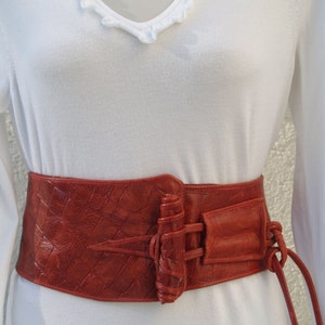 May include: A wide, red leather belt with a unique buckle design. The buckle is a large, textured piece of leather with a woven strap.