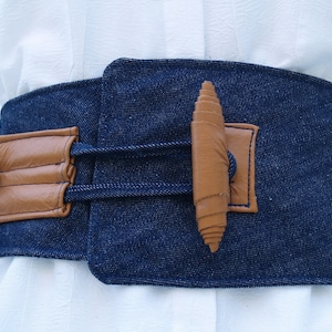 May include: A dark blue denim belt with a brown leather buckle and a brown leather strap with a decorative knot. The belt has a lace-up design on the side.