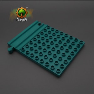 Könnte beinhalten: Eine teal-farbene Grundplatte für Bausteine mit einem Raster aus kreisförmigen Noppen. Die Grundplatte hat eine erhöhte Kante und ein kleines Verbindungsteil. Das Logo "Forgely" befindet sich in der oberen linken Ecke.