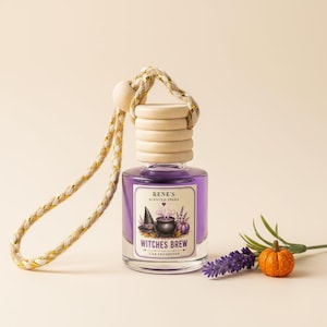 May include: A glass car freshener bottle filled with purple liquid, topped with a light wood cap and a braided gold and white hanging cord. The label reads RENE'S SCENTED SPARK WITCHES BREW CAR FRESHENER with a graphic of a witch hat and cauldron.