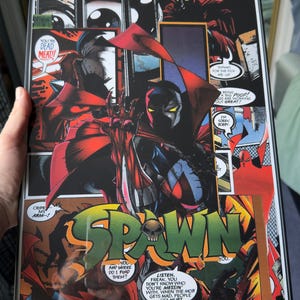 May include: Comic book art print featuring the character Spawn in a dynamic pose, with a red cape and black suit. The artwork includes speech bubbles and the title "SPAWN" in bold letters, with a skull graphic.