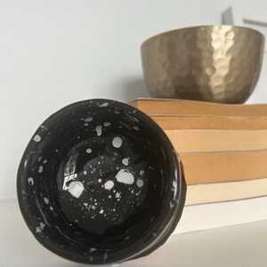 Peut inclure: Une tasse en céramique noire avec une finition brillante et un motif d'éclaboussures de peinture blanche à l'intérieur. La tasse est placée devant une pile de livres aux tons clairs et un bol doré métallique avec une texture martelée à l'extérieur.