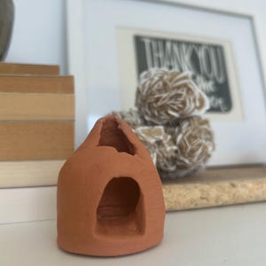 May include: A small, hand-sculpted terracotta-colored clay structure with a rounded dome shape, featuring a hollow interior and a single arched opening at the front. The surface has a matte, earthy texture with visible imperfections in the clay.