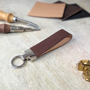May include: A brown leather keyring with a silver metal clasp and ring. The keyring is made from a folded strip of brown leather. Leather crafting tools and leather offcuts are in the background.
