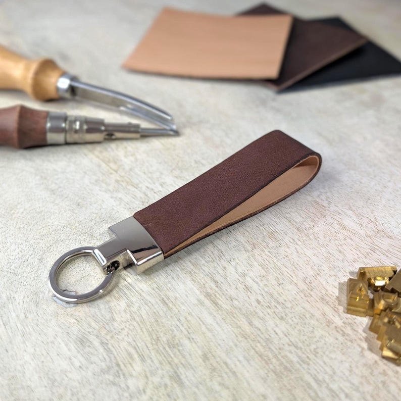 May include: A brown leather keyring with a silver metal clasp and ring. The keyring is made from a folded strip of brown leather. Leather crafting tools and leather offcuts are in the background.