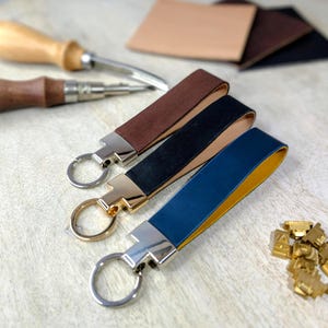 May include: Three leather keyrings in brown, black, and blue with silver and gold-toned hardware. The blue keyring has a yellow interior. Leathercraft tools and leather scraps are in the background. A handmade accessory.