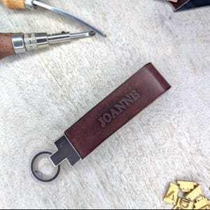 May include: A brown leather keychain with the name "JOANNE" embossed on it. The keychain has a silver-coloured metal clasp and key ring. Leatherworking tools are visible in the background.
