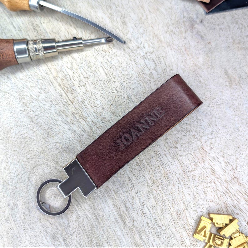 May include: A brown leather keychain with the name "JOANNE" embossed on it. The keychain has a silver-coloured metal clasp and key ring. Leatherworking tools are visible in the background.