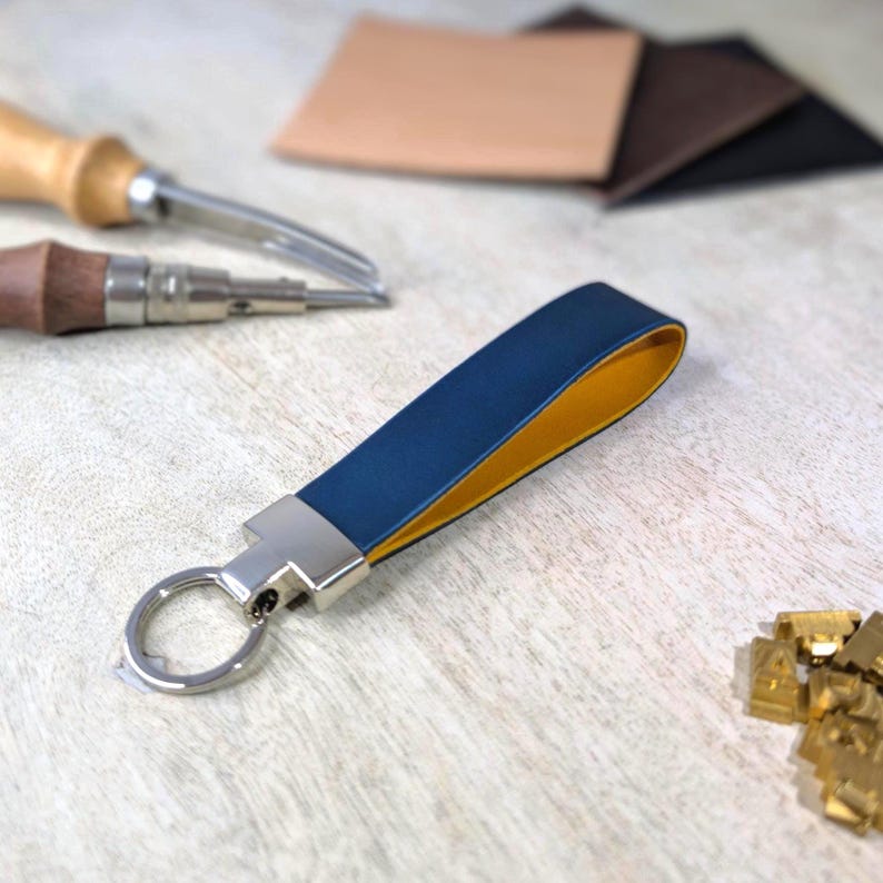 May include: A blue and yellow leather keyring with a silver metal ring and clasp. The keyring is on a light wooden surface, with leather crafting tools and leather scraps in the background.