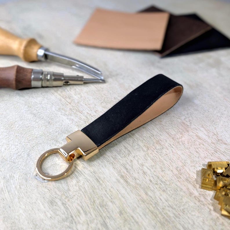 May include: Black leather keyring with a gold-coloured metal clasp and ring. The keyring has a loop design and is set against a light wood grain surface. Leather crafting tools and leather swatches are in the background.