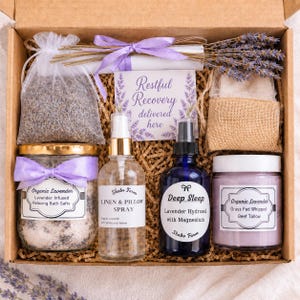 May include: A gift box containing a bag of dried lavender, a jar of bath salts with a purple ribbon, a clear spray bottle labeled Linen & Pillow Spray, a blue spray bottle labeled Deep Sleep, a jar of purple cream, and a card reading Restful Recovery.