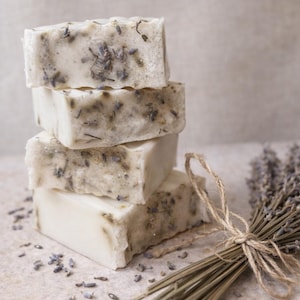 May include: A stack of four rectangular off-white soap bars embedded with dried purple lavender buds. Beside the soap stack lies a small bundle of dried lavender stems tied together with a thin piece of natural brown twine.