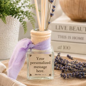 May include: A square glass reed diffuser bottle with a light wood collar and a tied lavender-colored frayed ribbon. The bottle features a label with the text 'Your personalized message here.' and 'Shabo Farm Benton, TN', containing wooden reeds and dried lavender sprigs.