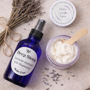 May include: A collection of lavender-scented products, including a blue spray bottle labeled "Deep Sleep Lavender Hydrosol with Magnesium," a jar of cream, a small jar with a lid, and a bundle of dried lavender stems. The products are arranged on a neutral surface.
