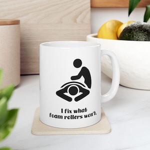 May include: White ceramic mug with a black graphic of a person giving a massage to another person. The text "I fix what foam rollers won't" is printed below the graphic. The mug is sitting on a beige coaster.