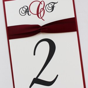 May include: White table number card with a burgundy ribbon tied in a bow. The card has a red monogram "ACF" and the number "2" in black script.