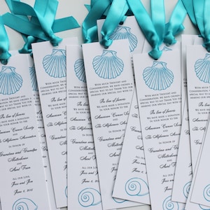 May include: A stack of white bookmark favors with a blue seashell design and a blue ribbon. The bookmarks have a message of thanks and a donation to the American Cancer Society and the Alpha Phi Omega Fraternity. The text reads "In lieu of favors, we have made a donation in your honor to the American Cancer Society and the Alpha Phi Omega Fraternity. In honor of Grandmother and Grandfather and Aunt Fran. All our love, Jess and Jon. June 8, 2019."