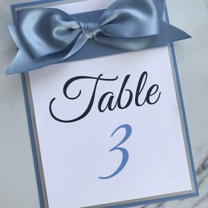 Puede incluir: Una tarjeta con el número de mesa con un lazo de satén azul claro. La tarjeta tiene un fondo blanco con la palabra "Table" en escritura negra y el número "3" en azul claro. La tarjeta está bordeada con plata y azul claro.