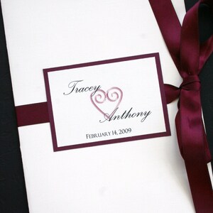 Puede incluir: Una invitación de boda blanca con una cinta color burdeos y un borde color burdeos alrededor de un rectángulo blanco. El rectángulo tiene los nombres "Tracey" y "Anthony" en letra cursiva con un símbolo de corazón rosa y la fecha "FEBRUARY 14, 2009".