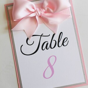 May include: A white card with a pink satin bow on top. The card has black text that says "Table 8" with the number 8 in pink.