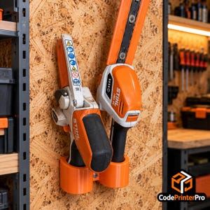 Op de afbeelding: Een oranje kunststof wandhouder met twee sleuven voor twee Stihl elektrische gereedschappen. Het linker gereedschap is een GTA 26 snoeizaag met veiligheidssymbolen, het rechter is een HSA 26 heggenschaar. Beide zijn wit en oranje van kleur.