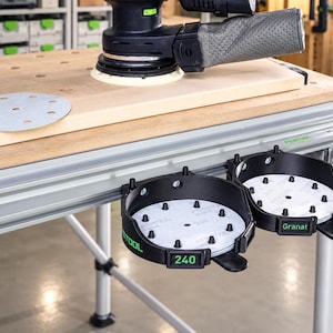 May include: A black Festool random orbital sander with a grey dust bag sits on a wooden workbench. Attached to the front of the workbench are two black circular storage holders containing white sanding discs labelled 240 and Granat.